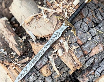 May include: A decorative sword with a silver blade featuring an etched leaf design. The hilt and crossguard are gold-colored, and the handle is wrapped in black material. The sword rests on a bed of wood.