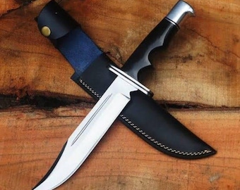 May include: A hunting knife with a polished steel blade and a black handle, resting in a black leather sheath. The sheath has a blue accent and is secured with a brass button. The knife is displayed on a wooden surface.