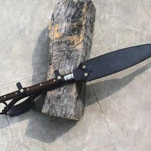 Puede incluir: Una lanza artesanal con un mango de madera oscura y una funda de cuero negro. La lanza presenta una punta de metal y detalles decorativos. La lanza descansa sobre un tronco de madera envejecida, mostrando su dise&ntilde;o r&uacute;stico.