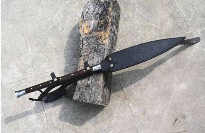 Puede incluir: Una lanza artesanal con un mango de madera oscura y una funda de cuero negro. La lanza presenta una punta de metal y detalles decorativos. La lanza descansa sobre un tronco de madera envejecida, mostrando su dise&ntilde;o r&uacute;stico.