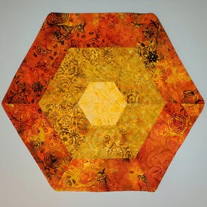 May include: A quilted, hexagonal placemat in shades of orange and yellow. The design features a central hexagon surrounded by layers of patterned fabric, with floral and abstract motifs. The outer layer is a vibrant orange, transitioning to yellow in the center.