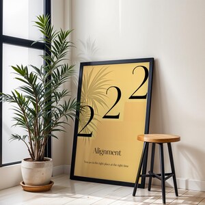 May include: A framed art print with the number "222" in black, and the word "Alignment" on a yellow background. A small wooden stool with black legs sits next to the print. A potted plant is in the foreground.