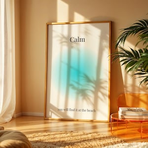 May include: A framed art print with the word "Calm" at the top and "you will find it at the beach" at the bottom. The artwork features a blue and white gradient design. The print is displayed in a room with natural light and a potted plant.