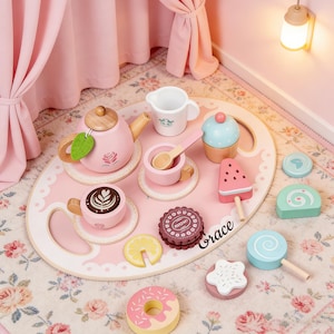 Può includere: Un set da tè rosa in legno con teiera, tazzine e vassoio. Il set include un cupcake, un ghiacciolo all'anguria e altre prelibatezze. Sul vassoio è scritto il nome "Grace". Il set è su un tappeto a fantasia floreale.