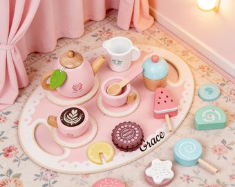 May include: A pink wooden tea set with a teapot, teacups, and a serving tray. The set includes a cupcake, a watermelon popsicle, and other treats. The tray has the name "Grace" written on it. The set is on a floral patterned rug.