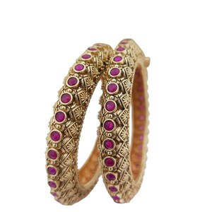 May include: Two gold bangles with detailed patterns and magenta gemstones. The bangles are set against a white backdrop, highlighting their ornate design and colour contrast. The jewellery appears to be made of metal.