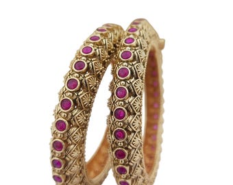 May include: Two gold-colored bangles with intricate patterns and embedded magenta-colored gemstones. The bangles are displayed against a white background, showcasing their ornate design and color contrast. The jewelry appears to be made of metal.