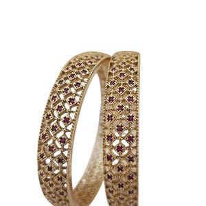 Op de afbeelding: Twee goudkleurige armbanden met ingewikkelde opengewerkte ontwerpen. Elke armband heeft een herhalend patroon van kleine, donkerrode edelstenen die in het opengewerkte werk zijn gezet. De armbanden worden gepresenteerd tegen een effen witte achtergrond.