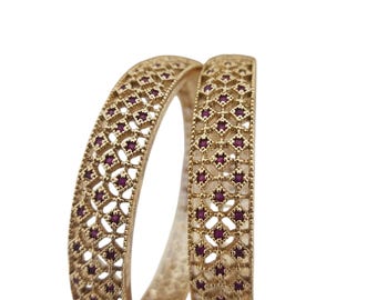 May include: Two gold-coloured bangles with intricate openwork designs. Each bangle features a repeating pattern of small, dark red gemstones set within the openwork. The bangles are displayed against a plain white background.