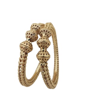 May include: Two gold-colored bangles. Each bangle features a series of decorative, spherical elements with intricate patterns. The bands have a textured, beaded design. The jewelry is set against a white background.