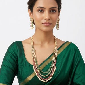 May include: A multi-layered gold necklace featuring red and green stones with small white pearl accents, paired with matching drop earrings. The jewelry set displays intricate metalwork and a tiered design that rests against a dark green fabric.