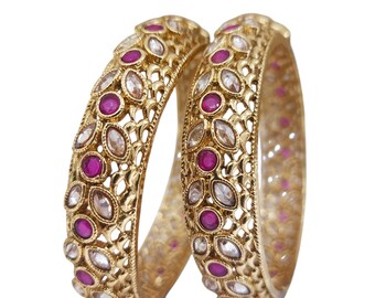 May include: Two gold-colored bangles with intricate openwork designs. Each bangle is adorned with alternating oval-shaped clear stones and round, deep pink gemstones. The bangles are set against a plain white background.