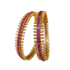 Op de afbeelding: Twee gouden armbanden met een herhalend patroon van vierkante, magenta edelstenen. De armbanden hebben een getextureerde gouden band met kleine, decoratieve gouden accenten tussen elke edelsteen. De achtergrond is effen wit.