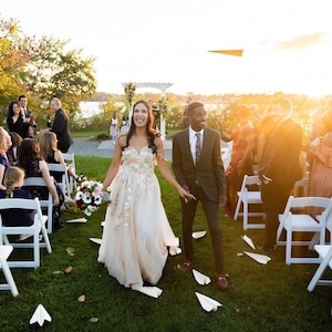 May include: A bride and groom walk down the aisle after their wedding ceremony, surrounded by guests throwing paper airplanes. The bride is wearing a white dress with floral embellishments and the groom is wearing a dark suit with a tie. The ceremony is taking place outdoors on a grassy lawn with white chairs set up for the guests.