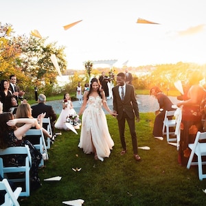 May include: A bride and groom walk down the aisle after their wedding ceremony, surrounded by guests throwing paper airplanes. The couple is smiling and holding hands, and the bride is wearing a white wedding dress with a long train. The groom is wearing a dark suit and tie. The ceremony is taking place outdoors in a grassy area with a white arbor in the background.