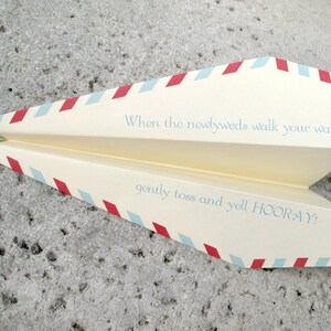 May include: A paper airplane with red and blue stripes, folded in half. The text on the airplane reads "When the newlyweds walk your way... gently toss and yell HOORAY!"