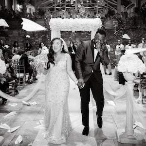 May include: A black and white photo of a bride and groom walking down the aisle after their wedding ceremony. Guests are throwing paper airplanes at them as they exit the ceremony space.