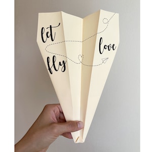 May include: A beige paper airplane with the text "let love fly" written on it. The airplane is folded in the shape of a heart with a dashed line connecting the two words.