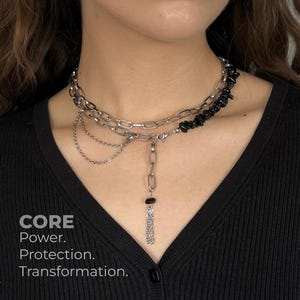 May include: A layered silver-tone necklace with a chain design, featuring a dangling pendant with a black stone and fringe. The necklace also includes a section of black stones. The image includes the text: CORE. Power. Protection. Transformation.