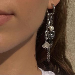 May include: Silver dangle earrings with multiple chains, a pearl, and a small, irregular-shaped stone. The earrings are attached to a silver hoop. The design is modern and features a mix of textures and shapes.