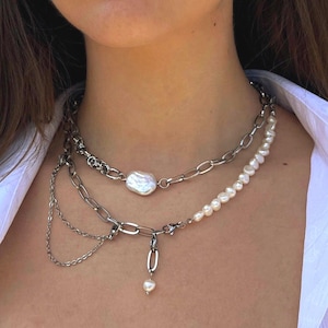 Puede incluir: Un collar de varias capas con un colgante de perla grande e irregular y una cadena con una pequeña gota de perla. El collar tiene una cadena plateada con una mezcla de eslabones grandes y pequeños y una hebra de pequeñas perlas.