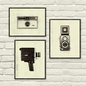 May include: Three black framed prints of vintage cameras on a white brick wall. The prints feature a black and silver 35mm camera, a black and silver rangefinder camera, and a black and silver film camera.