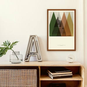 May include: A framed art print featuring a mountain landscape with geometric shapes in green, orange, and brown. The print is displayed on a wooden cabinet with a decorative letter "A" and a small clock. A glass vase with a green plant is also on the cabinet.