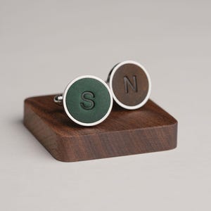 Może przedstawiać: Para spersonalizowanych spinek do mankietów ze srebrną obwódką. Jedna ma ciemnozielony środek z literą "S", druga brązowy środek z literą "N". Prezentowane są na małym, ciemnym drewnianym bloku.