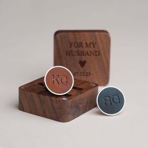 May include: Personalised cufflinks with initials and a wooden box. One cufflink has "KC" on a brown leather surface, the other has "RG" on a dark blue surface. The box is engraved with "FOR MY HUSBAND" and a date.