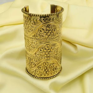 May include: A gold-colored cuff bracelet with intricate floral and circular patterns. The wide bracelet has a textured surface and a beaded edge. The open design allows for easy wear. The jewelry piece is displayed on a pale yellow fabric.