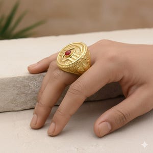 May include: A gold-colored ring with a central red stone is displayed. The ring has an intricate design with a textured surface and a circular shape. The ring is worn on a finger, showcasing its details and craftsmanship.