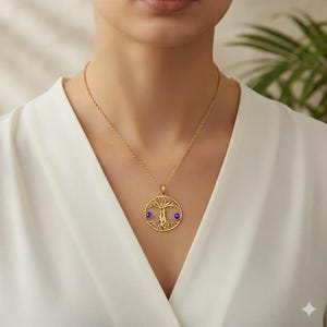 Peut inclure: Un collier pendentif doré avec un motif d'arbre de vie. Le pendentif comporte deux petites pierres précieuses rondes et violettes. Le collier est porté sur un haut blanc.