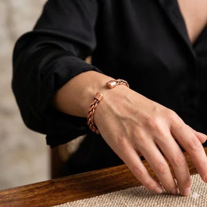 May include: A copper-colored bracelet with a braided design. The bracelet has a rectangular clasp on each end. The jewelry is worn on a person's wrist, against a black shirt.