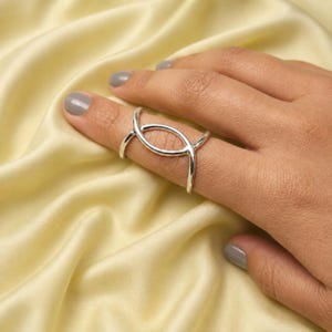 Puede incluir: Un anillo de plata pulida con un diseño de doble banda que se cruza para formar una forma ovalada abierta y alargada. La banda se envuelve alrededor del dedo, creando una estructura metálica continua y suave con un acabado reflectante.