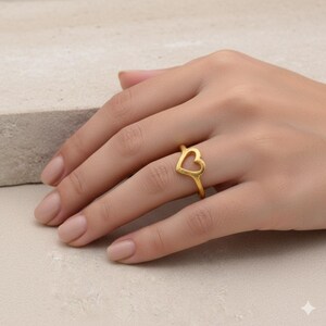 May include: A gold-colored ring featuring a heart-shaped design. The ring is worn on a finger, showcasing its delicate and romantic aesthetic. The ring's simple yet elegant design makes it a versatile accessory.