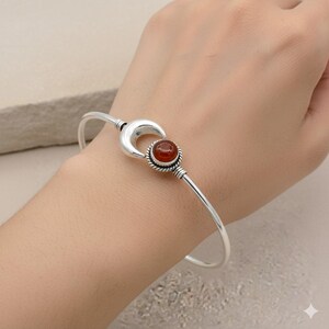 Könnte beinhalten: Ein silberfarbenes Armreif-Armband mit einem Halbmond-Design und einem zentralen, runden, rot-orangen Edelstein. Der Edelstein ist von einem verdrehten Metallrand umgeben. Das Armband ist ein schlichtes, elegantes Design.