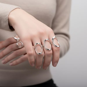 以下が含まれることがあります： 手には、オープンオーバルデザインのシルバーカラーのリングがはめられています。 リングは複数の指に着用され、モダンでミニマリストな美しさを強調しています。 リングは光沢のある金属で作られています。