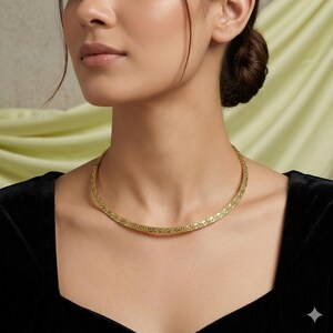 May include: A gold-colored necklace with a textured surface. The necklace is a choker style, sitting close to the neck. The background is a light green color.