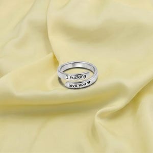 Puede incluir: Un anillo abierto de tono plateado con diseño cruzado. La banda metálica tiene grabado el texto en negro "I fucking" en la parte superior y "love you" seguido de un pequeño símbolo de corazón negro en la parte inferior que se superpone.