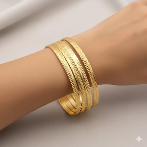 Puede incluir: Un conjunto de brazaletes dorados con un patrón de espiga texturizado. Las pulseras están apiladas en una muñeca, mostrando su diseño y brillo. Las joyas se exhiben sobre un fondo neutro, resaltando el color dorado.