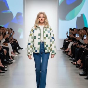 May include: A woman wearing a quilted jacket with a square pattern in shades of green and blue, paired with blue jeans and a white top. The jacket has a green trim. The model is walking on a runway with an audience on either side.