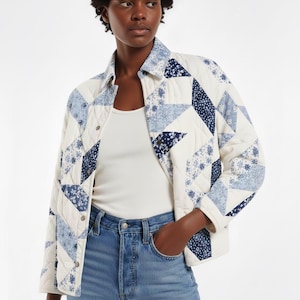 May include: A cream-coloured quilted jacket with a patchwork design. It features blue floral and geometric patterns. The jacket has a button-up front, a collar, and long sleeves. It is worn with a white top and blue jeans.