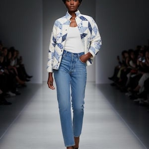 May include: A model wearing a white top, light blue jeans, and a quilted jacket with a blue and white patchwork design. The jacket has a stand-up collar and button closures. The jeans are high-waisted with a straight leg. The model is walking on a runway.