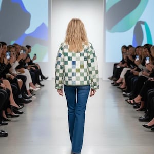 May include: A model walks a runway wearing a patchwork jacket with green, blue, and white squares, paired with blue jeans. The jacket has a quilted appearance. Spectators are seated on both sides of the runway, taking photos.