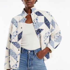 May include: A quilted jacket in a patchwork design. The jacket features a cream-colored base with blue and navy geometric patterns. It has a stand-up collar, snap closures, and long sleeves. The model is wearing a white tank top and blue jeans.
