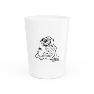 May include: White shot glass featuring a black and white cartoon illustration of a creature with two eyes and a spider hanging from a string. The creature has a patterned body and a spider hanging from a string.