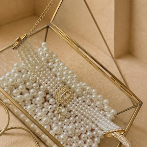 Peut inclure: Une boîte rectangulaire en verre avec un cadre en métal doré contenant un bracelet de perles à plusieurs rangs. Le bracelet présente des rangées de fausses perles blanches et un fermoir rectangulaire doré orné de cristaux et d'une breloque.
