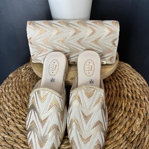 Könnte beinhalten: Ein Set aus cremefarbenen Schuhen und einer Clutch mit goldenem Zickzackmuster. Die Schuhe sind Mules mit spitzer Zehenpartie. Das Set wird auf einer gewebten Oberfläche präsentiert. Die Schuhe sind Größe 39, was etwa 24,5 cm entspricht.
