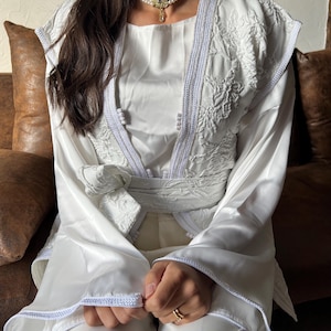 May include: A white satin-like jacket featuring intricate tonal embroidery on the front panels and shoulders, finished with a braided trim along the edges. The garment includes a matching fabric tie belt at the waist and a multi-strand pearl choker.