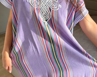 May include: A light purple kaftan featuring vertical stripes in shades of red, blue, yellow, and white. The garment has short sleeves and a V-neckline adorned with intricate white cord embroidery in a decorative swirling pattern.